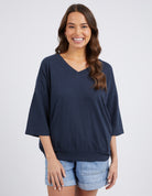 A woman modeling a dark navy blue V-neck knit top with wide three-quarter sleeves, paired with light blue denim shorts, against a light gray studio background.