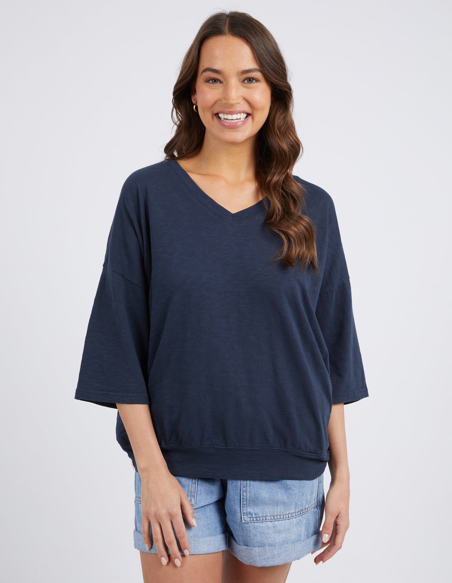 A woman modeling a dark navy blue V-neck knit top with wide three-quarter sleeves, paired with light blue denim shorts, against a light gray studio background.