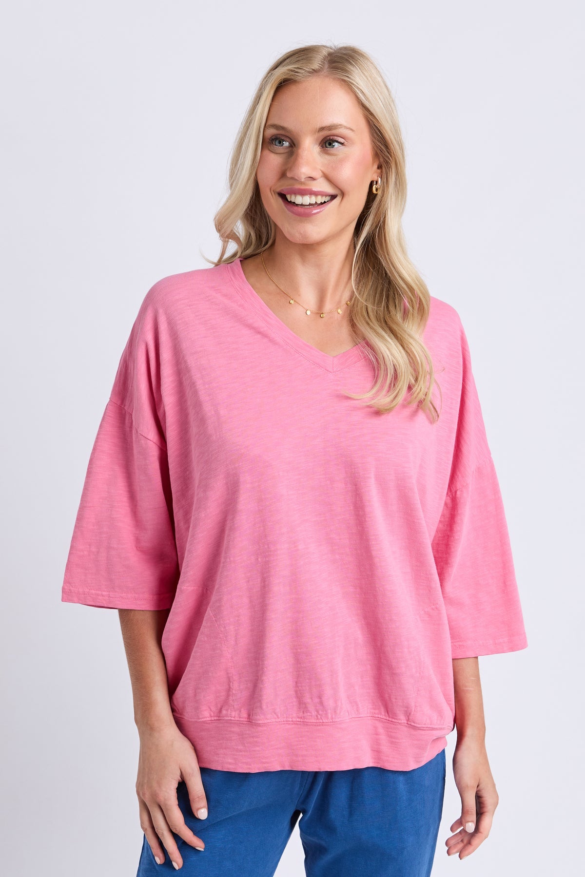 A smiling blonde woman wearing a pink V-neck knit top with three-quarter sleeves and blue pants, standing against a light gray studio background.