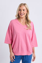 A smiling blonde woman wearing a pink V-neck knit top with three-quarter sleeves and blue pants, standing against a light gray studio background.