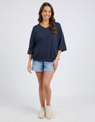 A woman modeling a navy blue oversized V-neck t-shirt with three-quarter sleeves, paired with light-wash denim shorts and beige sneakers, against a light gray studio background.