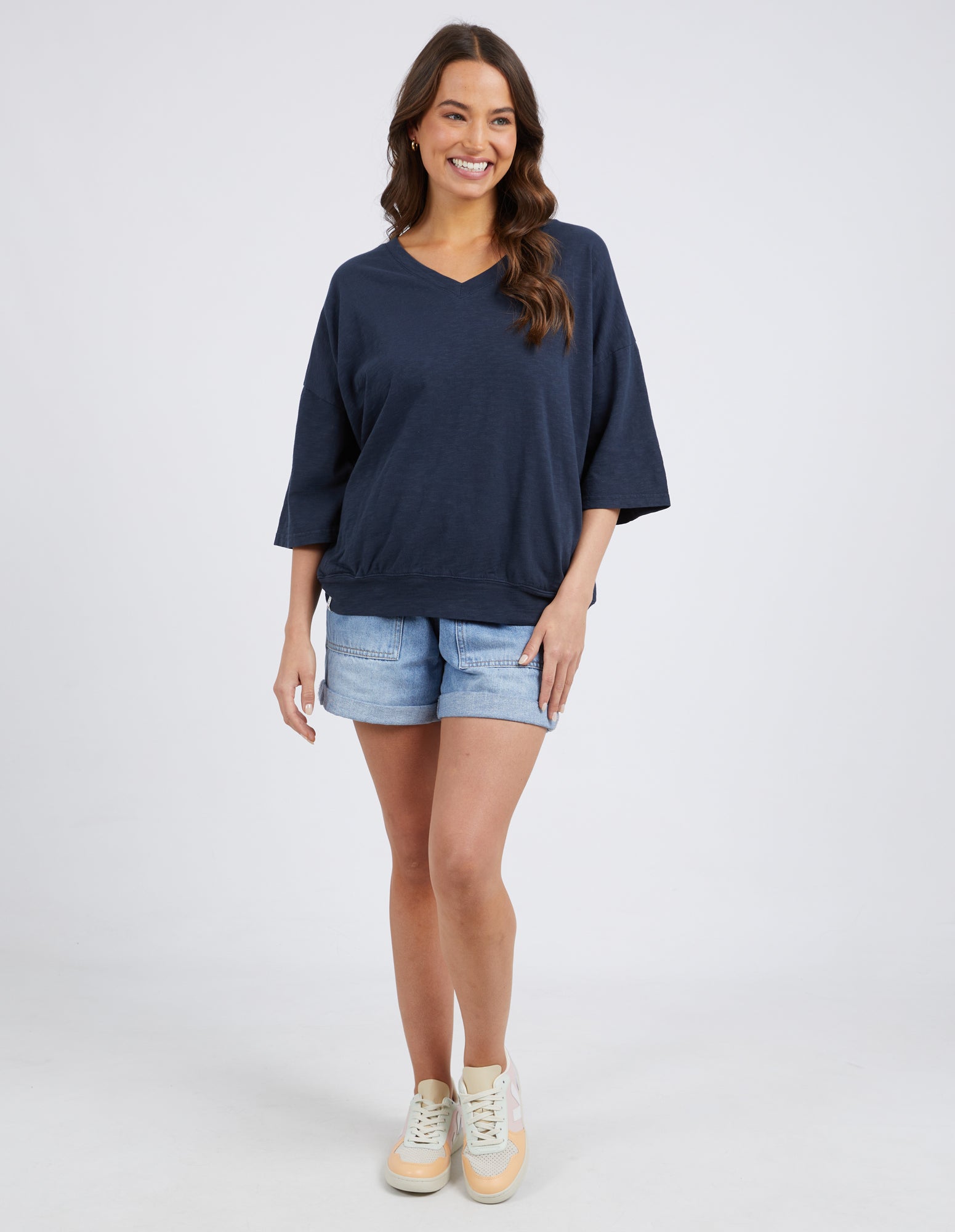 A woman modeling a navy blue oversized V-neck t-shirt with three-quarter sleeves, paired with light-wash denim shorts and beige sneakers, against a light gray studio background.
