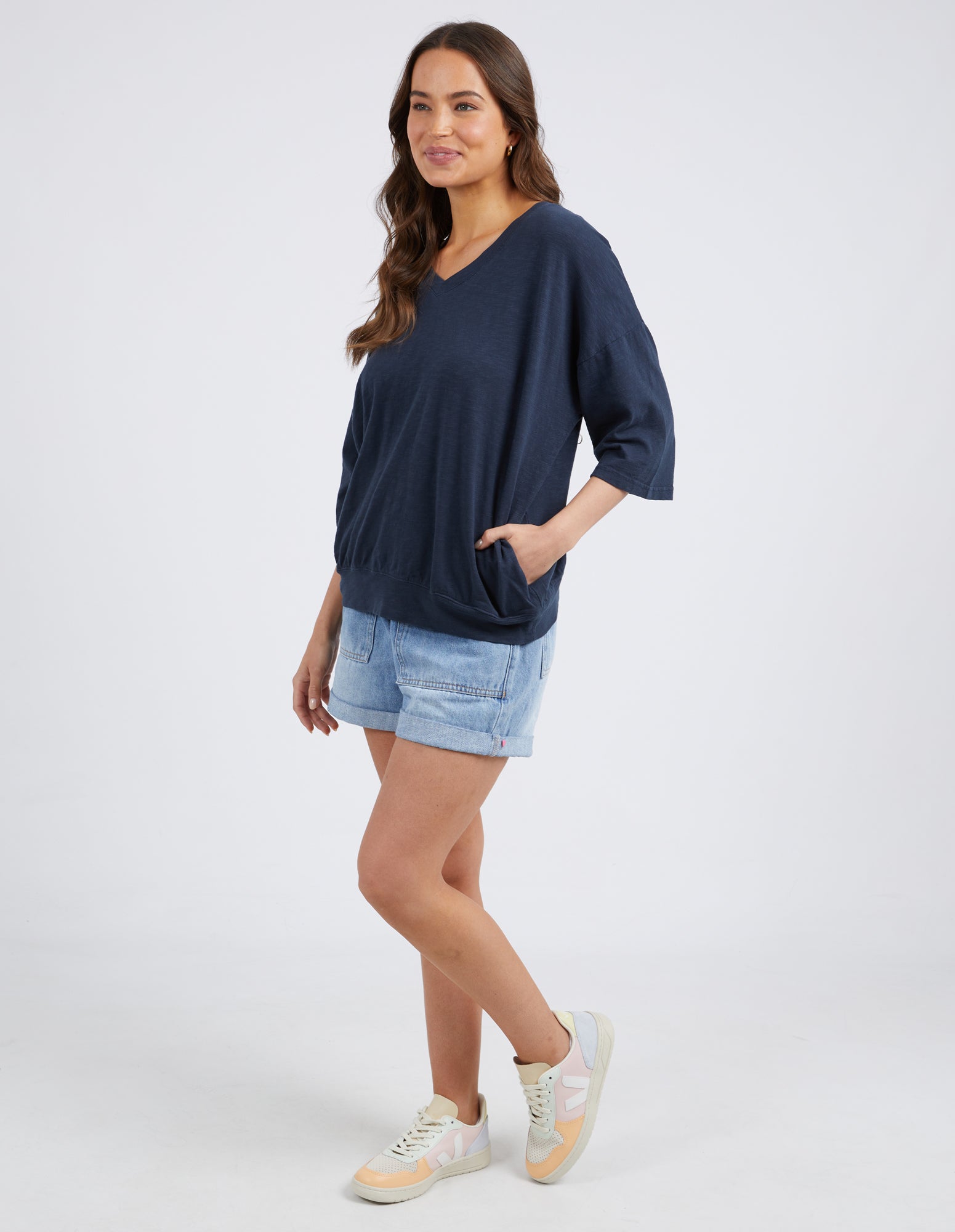 A woman modeling a loose-fitting navy blue V-neck top with three-quarter sleeves, paired with light denim shorts and white sneakers, against a light gray studio background.