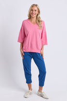 A woman modeling a pink V-neck short-sleeve top and blue cropped pants with white sneakers, standing against a light gray studio background.