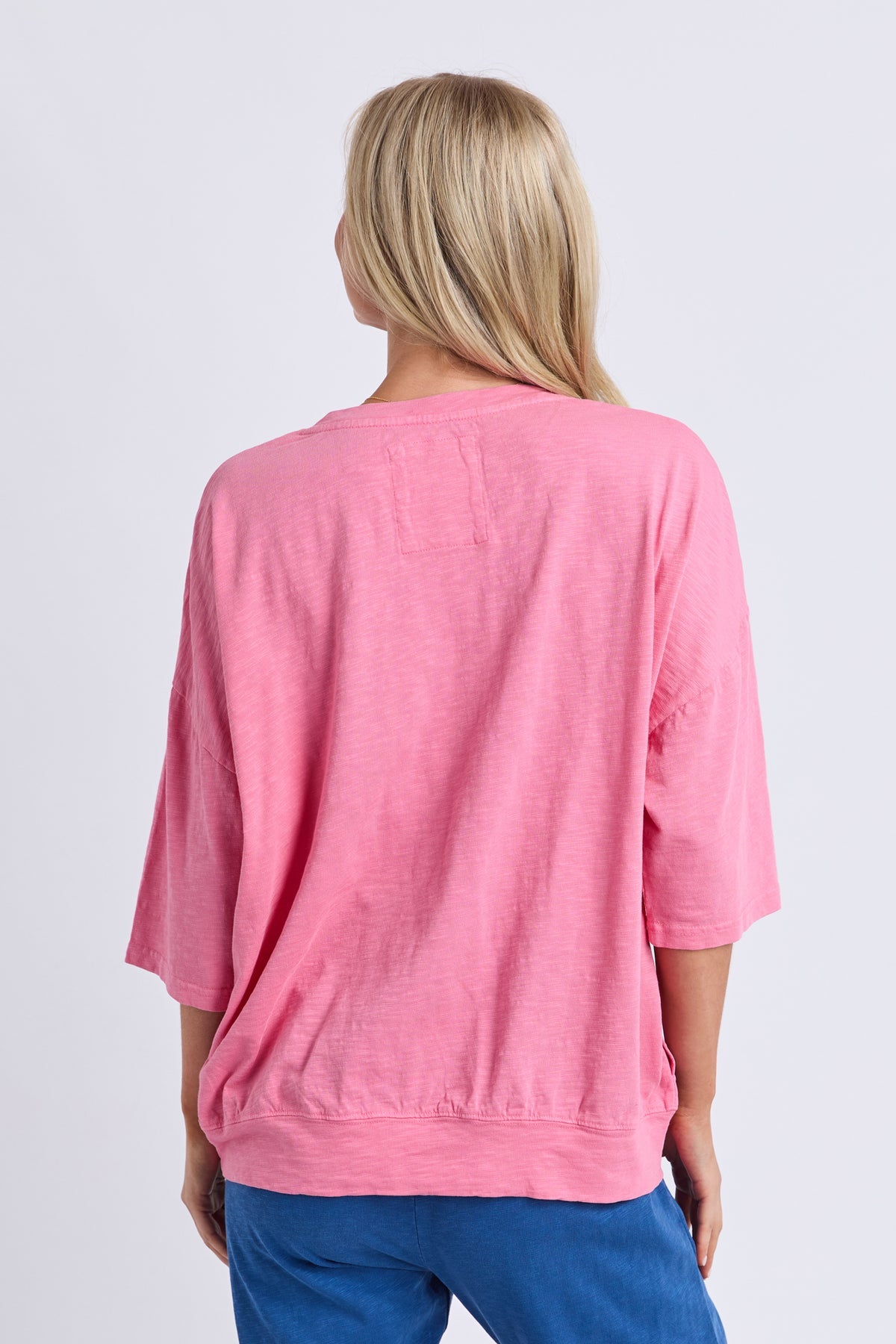 A woman viewed from the back wearing a pink short-sleeve top and blue pants against a light gray studio background.