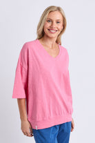 A smiling blonde woman wearing a pink V-neck knit top with three-quarter sleeves and blue pants, standing against a light gray studio background.