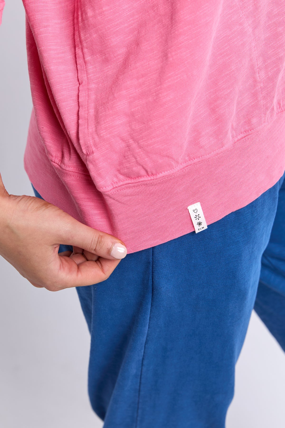 A close-up of a person wearing a pink heathered sweatshirt and blue sweatpants, with a hand pulling slightly away from the hem of the sweatshirt to show a small white brand tag.