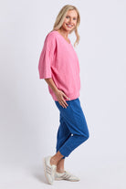 A woman modeling a pink oversized short-sleeve top and blue jogger pants with white sneakers, standing against a light gray studio background.