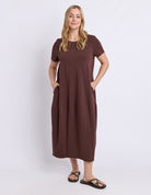 A woman modeling a short-sleeve brown midi dress with side pockets, paired with black sandals, against a plain light gray studio background.