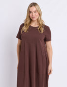 A woman modeling a short-sleeve, crew-neck dress in a solid dark brown color, photographed against a light gray studio background.