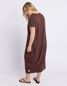 A woman wearing a brown short-sleeve midi dress with large patch pockets, shown from a side-back angle against a light gray studio background.