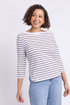 A smiling woman with curly brown hair wearing a white three-quarter sleeve top with thin dark brown horizontal stripes and light blue jeans, standing against a light gray studio background.