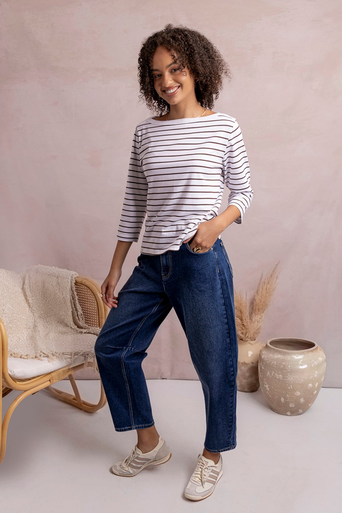 A woman modeling a white long-sleeve top with thin navy horizontal stripes, paired with dark blue cropped jeans and beige sneakers, standing in a styled room with a pink wall, a chair, and decorative vases.