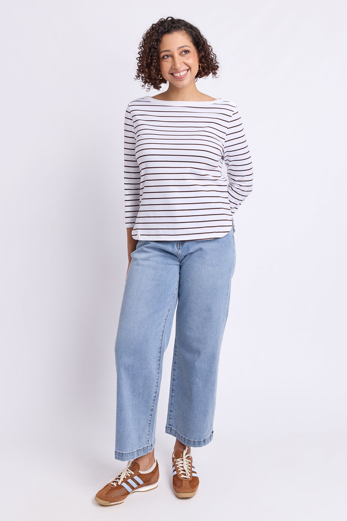 A woman modeling a white long-sleeve top with thin dark horizontal stripes, paired with light blue wide-leg jeans and brown sneakers, against a plain white studio background.