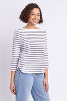 A woman with curly brown hair wearing a white three-quarter sleeve top with thin dark brown horizontal stripes and light blue jeans, standing against a light gray studio background.