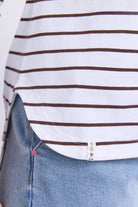 Close-up of a white short-sleeve top with thin dark brown horizontal stripes, worn over light blue denim jeans. A small sewn label is visible near the hem.