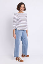 A woman modeling a white long-sleeve top with thin dark horizontal stripes, paired with light blue wide-leg jeans and brown sneakers, against a plain white studio background.