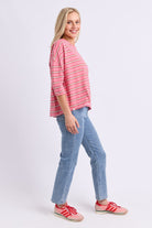 A woman modeling a pink and white striped three-quarter sleeve top, paired with light blue jeans and pink sneakers, against a light gray studio background.