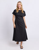 A woman modeling a black midi dress with short sleeves and a boat neckline, standing against a light gray studio background.