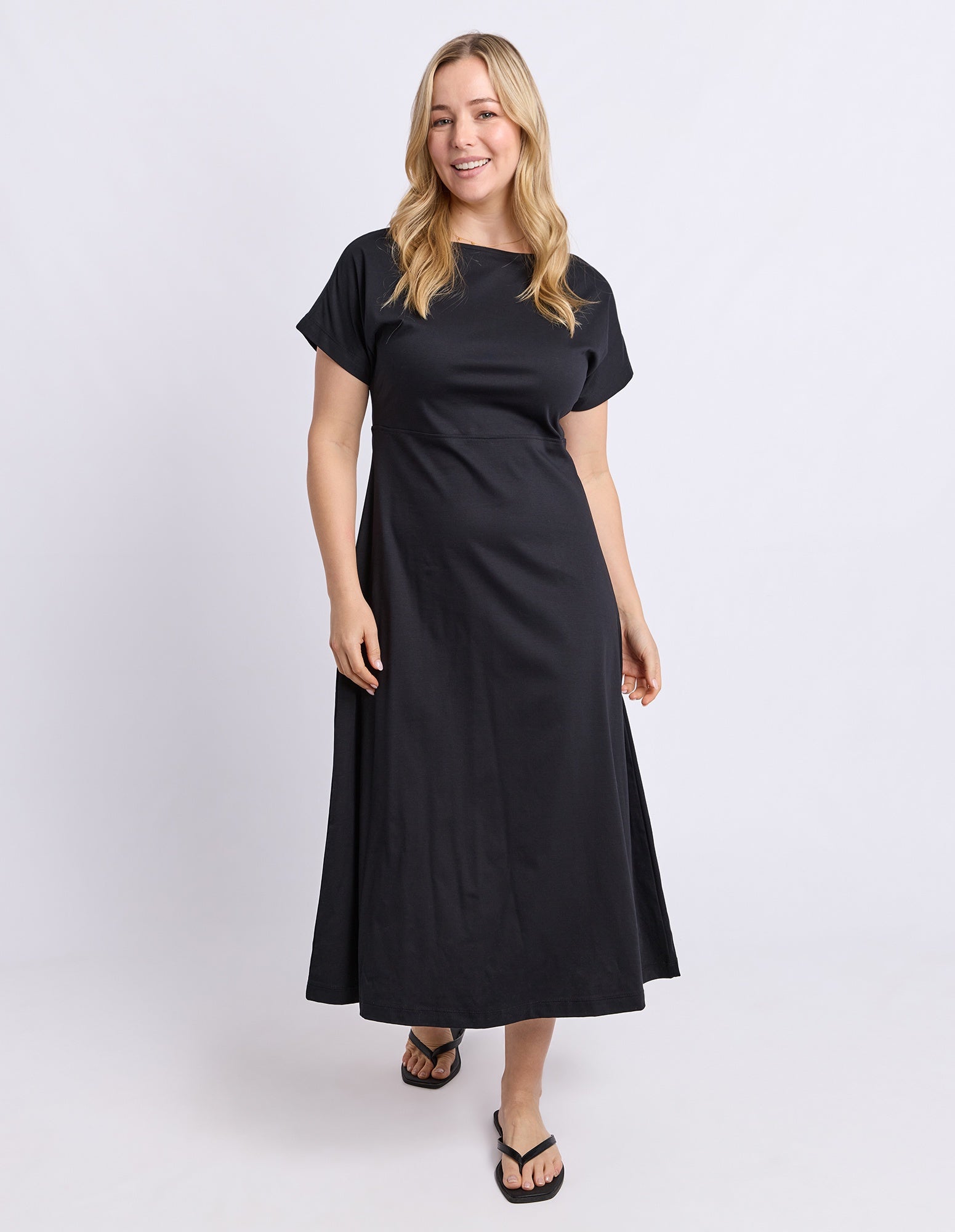 A woman modeling a black midi dress with short sleeves and a boat neckline, standing against a light gray studio background.