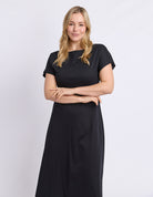 A woman with blonde hair wearing a black short-sleeve maxi dress, standing against a light gray studio background with her arms crossed.