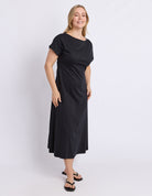 A woman modeling a black short-sleeve midi dress with a boat neckline, photographed against a plain light gray studio background.
