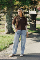 Woman wearing a brown sweater and light blue jeans standing on a path with trees in the background