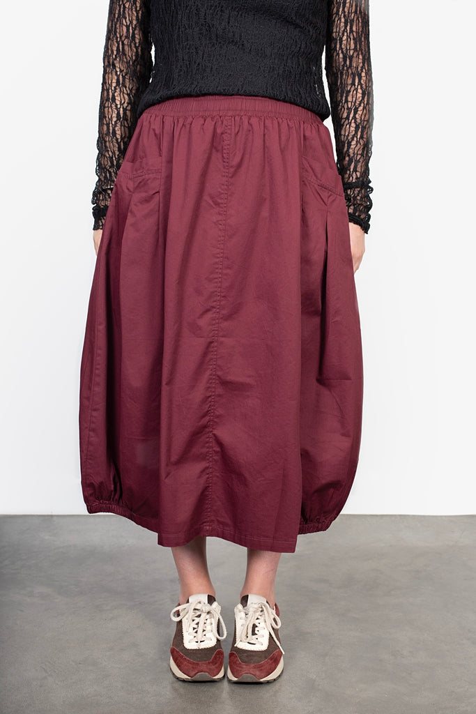 Person wearing a burgundy skirt with a black top and red shoes on a plain background