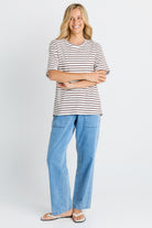 A woman modeling a short-sleeve crewneck t-shirt with thin brown and white horizontal stripes, paired with light blue wide-leg jeans and beige sandals, against a plain light gray studio background.