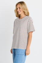 A woman wearing a short-sleeve crewneck t-shirt with thin brown horizontal stripes on an off-white base, paired with light blue jeans, photographed against a light gray studio background.