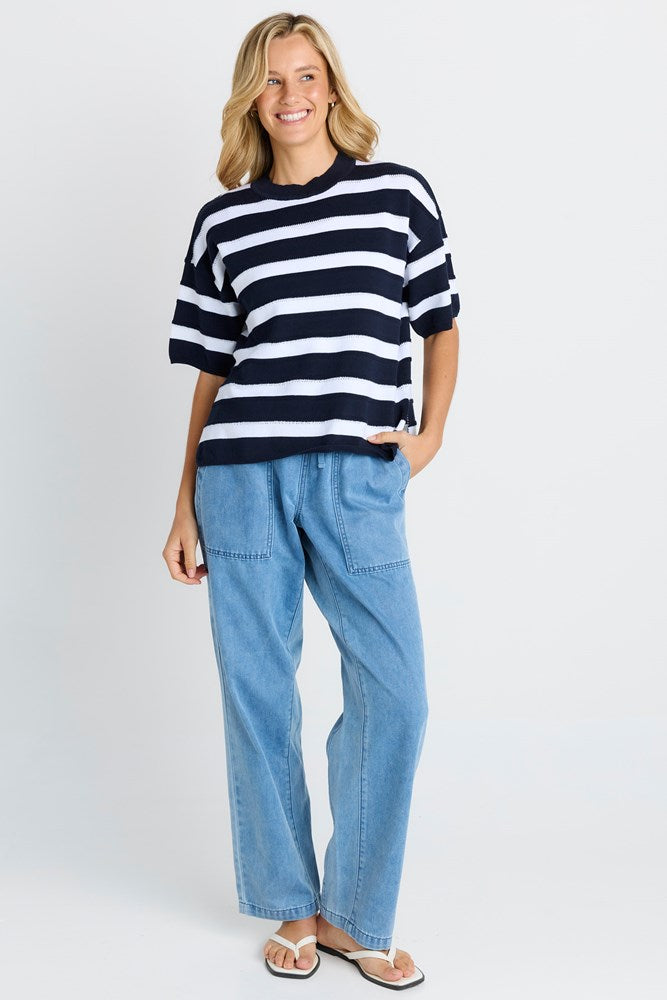 A woman modeling a navy and white horizontally striped short-sleeve knit top with light blue wide-leg jeans and white slide sandals, standing against a light gray studio background.