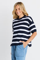 A woman wearing a navy blue and white horizontally striped short-sleeve knit top with light blue jeans, standing against a light gray studio background.