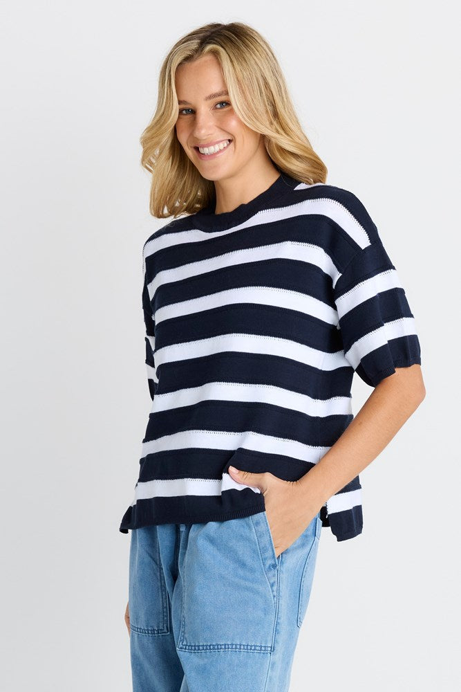 A woman wearing a navy blue and white horizontally striped short-sleeve knit top with light blue jeans, standing against a light gray studio background.