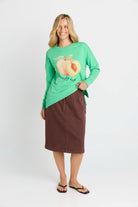 Woman wearing a green long-sleeve shirt with a peach graphic and brown skirt on a white background