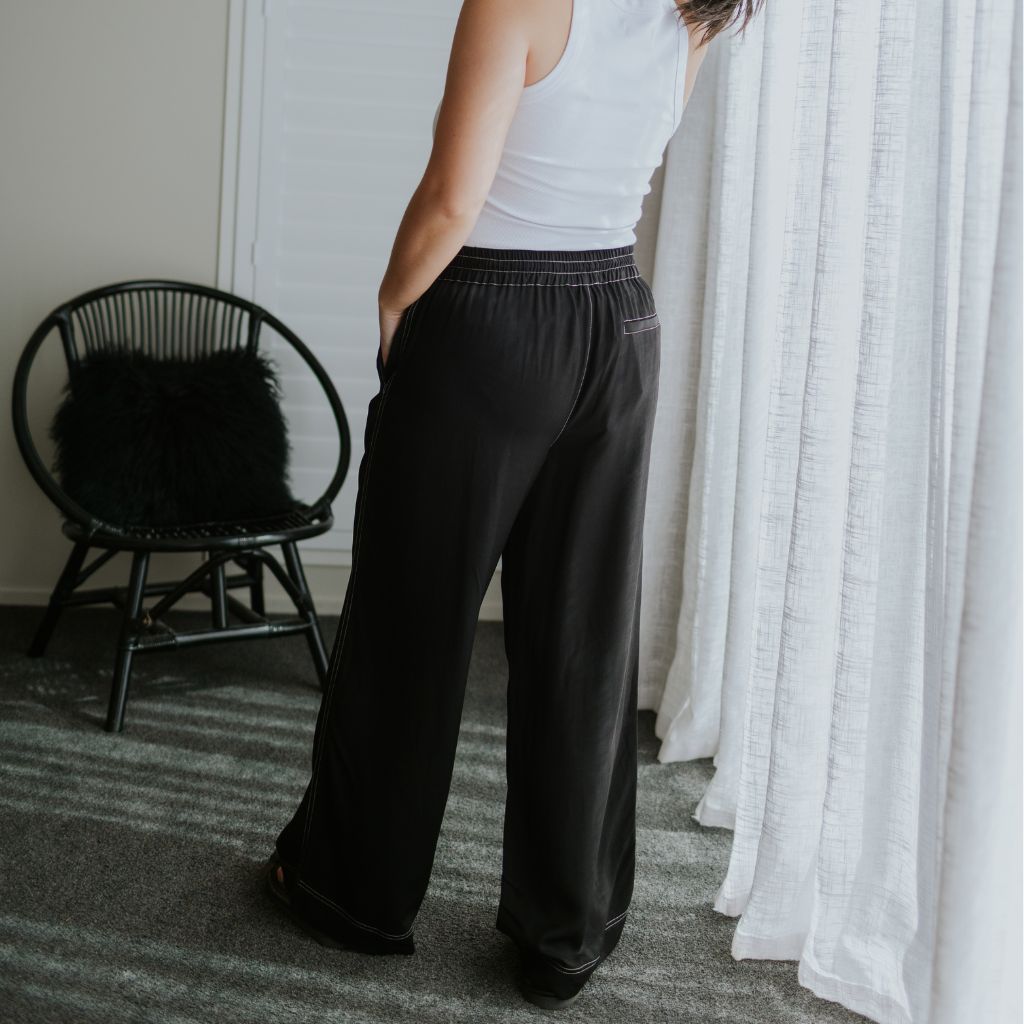 A person wearing black wide-leg pants with an elastic waistband, paired with a white tank top, standing in a room with white curtains and a black chair.