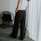 A person wearing black wide-leg pants with an elastic waistband, paired with a white tank top, standing in a room with white curtains and a black chair.