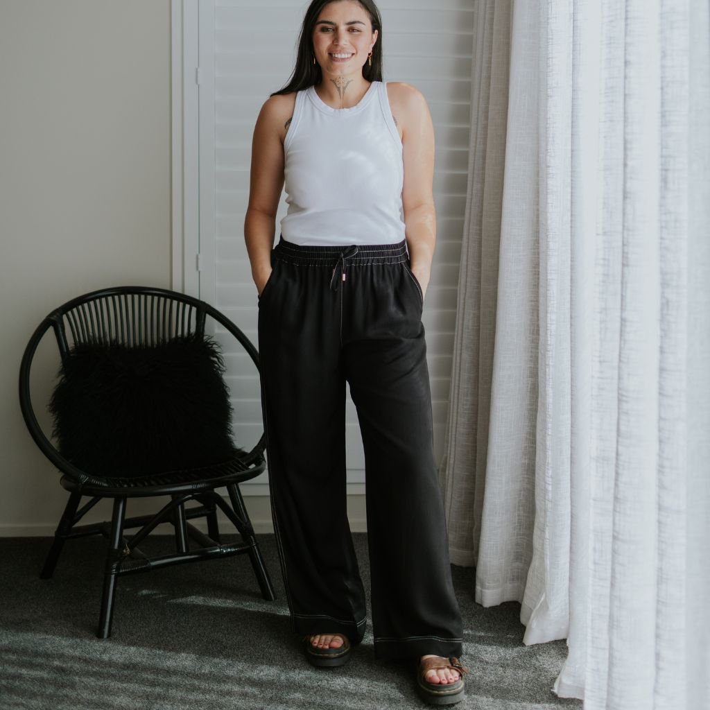 A woman stands indoors wearing loose black drawstring pants and a white sleeveless top, with a black chair and white curtains in a neutral room.