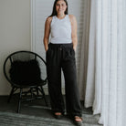 A woman stands indoors wearing loose black drawstring pants and a white sleeveless top, with a black chair and white curtains in a neutral room.