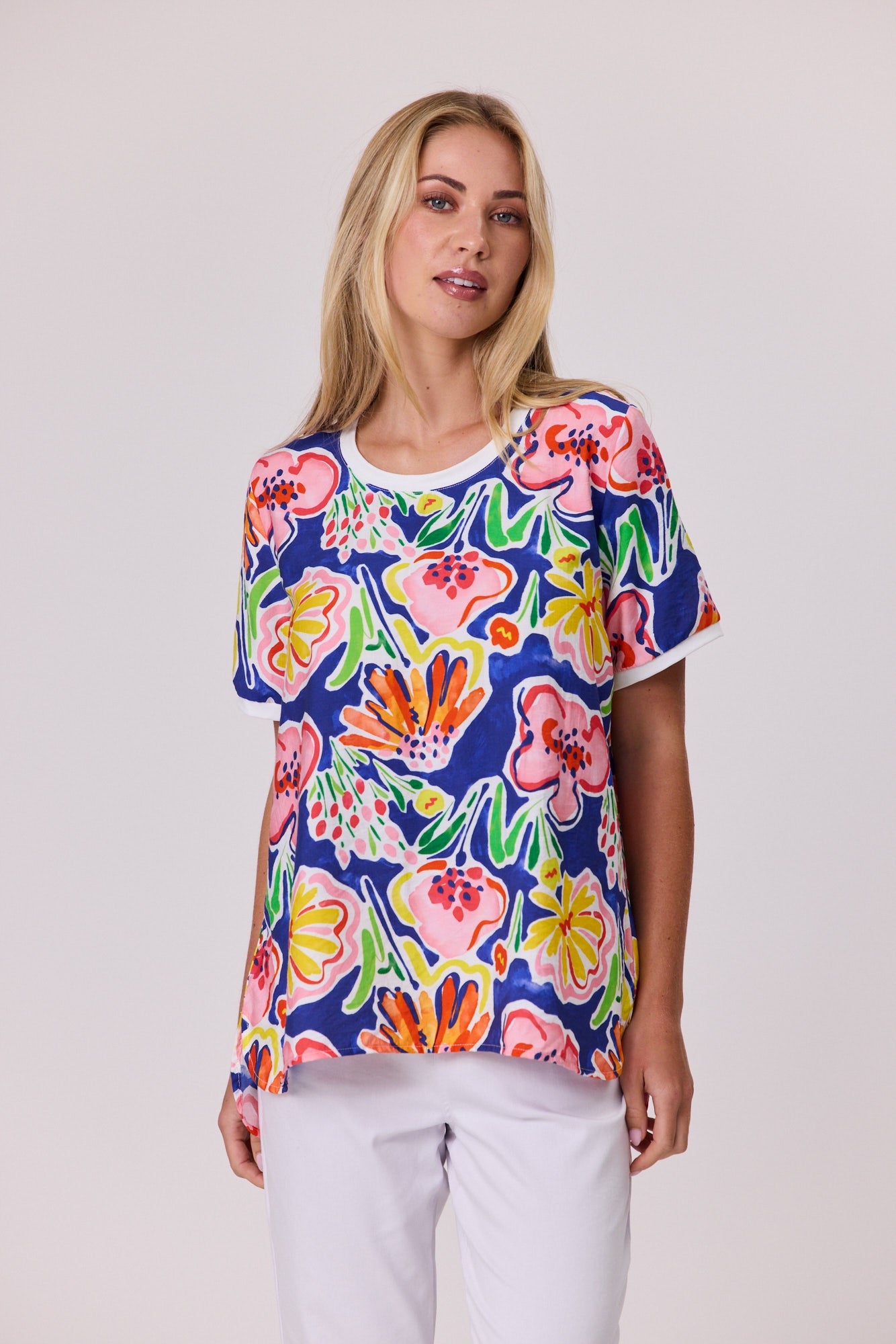A woman modeling a short-sleeve crewneck top with a vibrant multicolor floral print on a navy blue background, paired with white pants, against a solid light gray studio background.