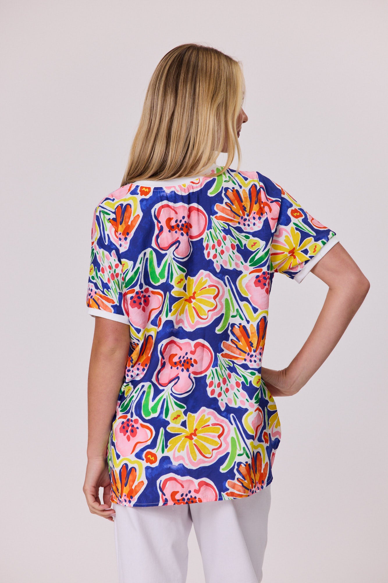 A woman shown from the back wearing a short-sleeve scrub top with a vibrant multicolor floral print on a navy blue background, paired with white pants, against a light gray studio backdrop.