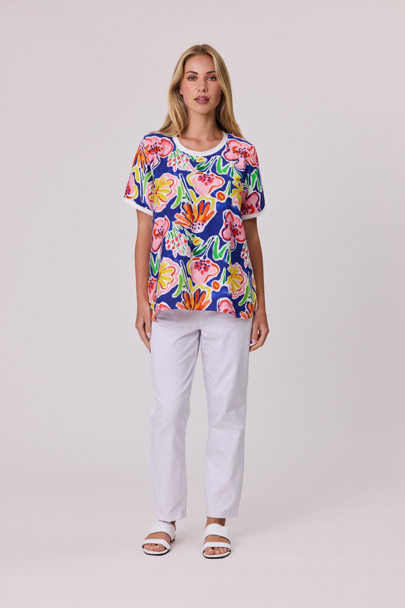A woman modeling a short-sleeve crewneck top with a vibrant multicolor floral print on a navy blue background, paired with white pants and white slide sandals, against a light gray studio background.