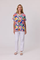 A woman modeling a short-sleeve crewneck top with a vibrant multicolor floral print on a navy blue background, paired with white pants and white slide sandals, against a light gray studio background.