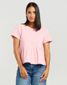 A woman modeling a light pink short-sleeve top with a relaxed, slightly ruffled hem, paired with blue jeans, against a solid light gray background.