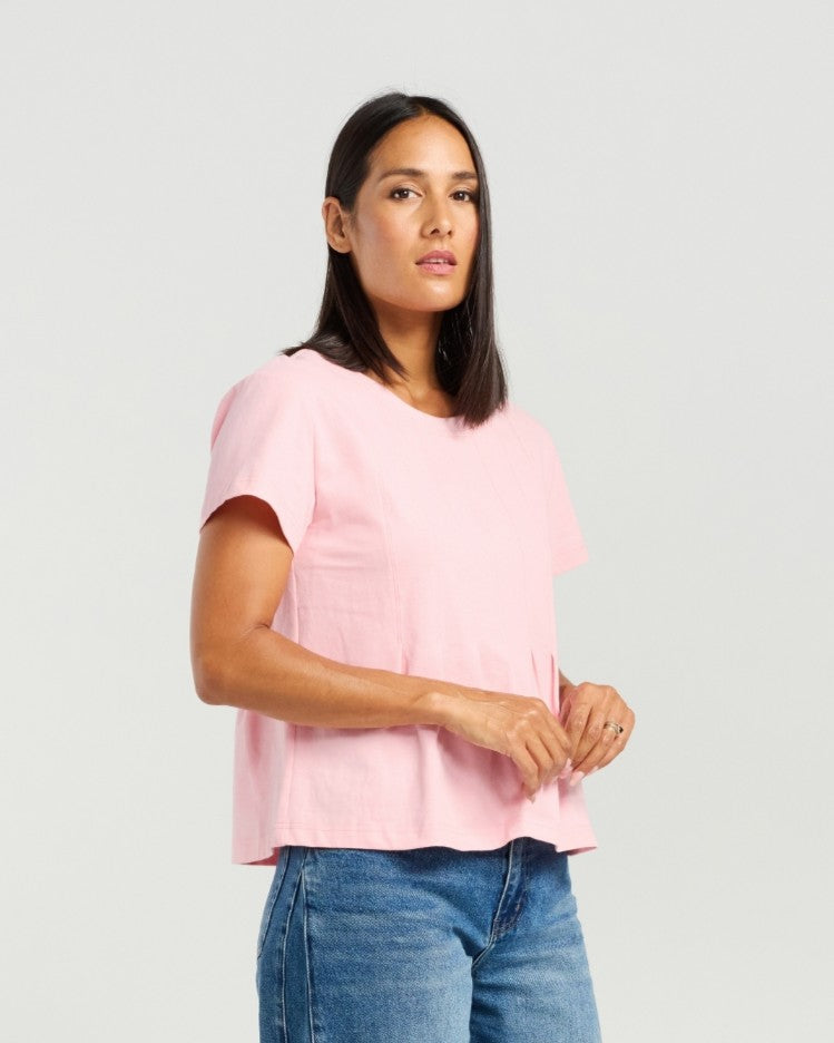 A woman modeling a light pink short-sleeve top with a relaxed, slightly ruffled hem, paired with blue jeans, against a solid light gray background.