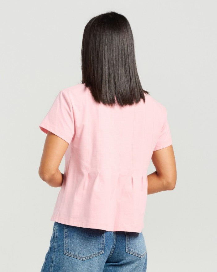 A woman modeling a light pink short-sleeve top with a relaxed, slightly ruffled hem, paired with blue jeans, against a solid light gray background.