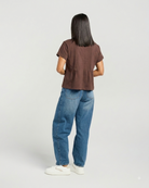A woman shown from the back wearing a brown short-sleeve cropped top, blue jeans, and white sneakers against a light gray studio background.
