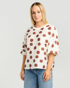 A woman modeling a cream-colored short-sleeve top with large brown polka dots, paired with blue jeans, against a light gray studio background.