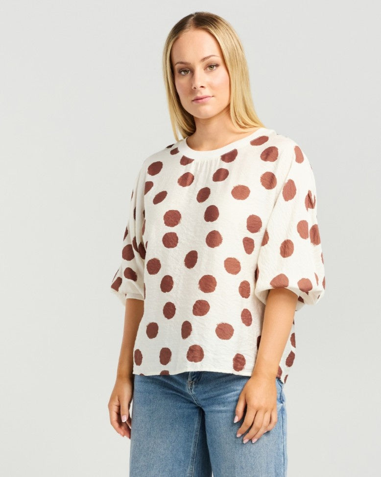 A woman modeling a cream-colored short-sleeve top with large brown polka dots, paired with blue jeans, against a light gray studio background.