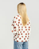 A woman with long blonde hair is shown from behind wearing a cream-colored short-sleeve top with large brown polka dots, paired with light blue jeans, against a light gray studio background.
