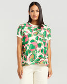 A woman modeling a short-sleeve crewneck top with a vibrant green and pink abstract floral pattern on an off-white background, paired with cream-colored pants, against a solid light gray studio background.